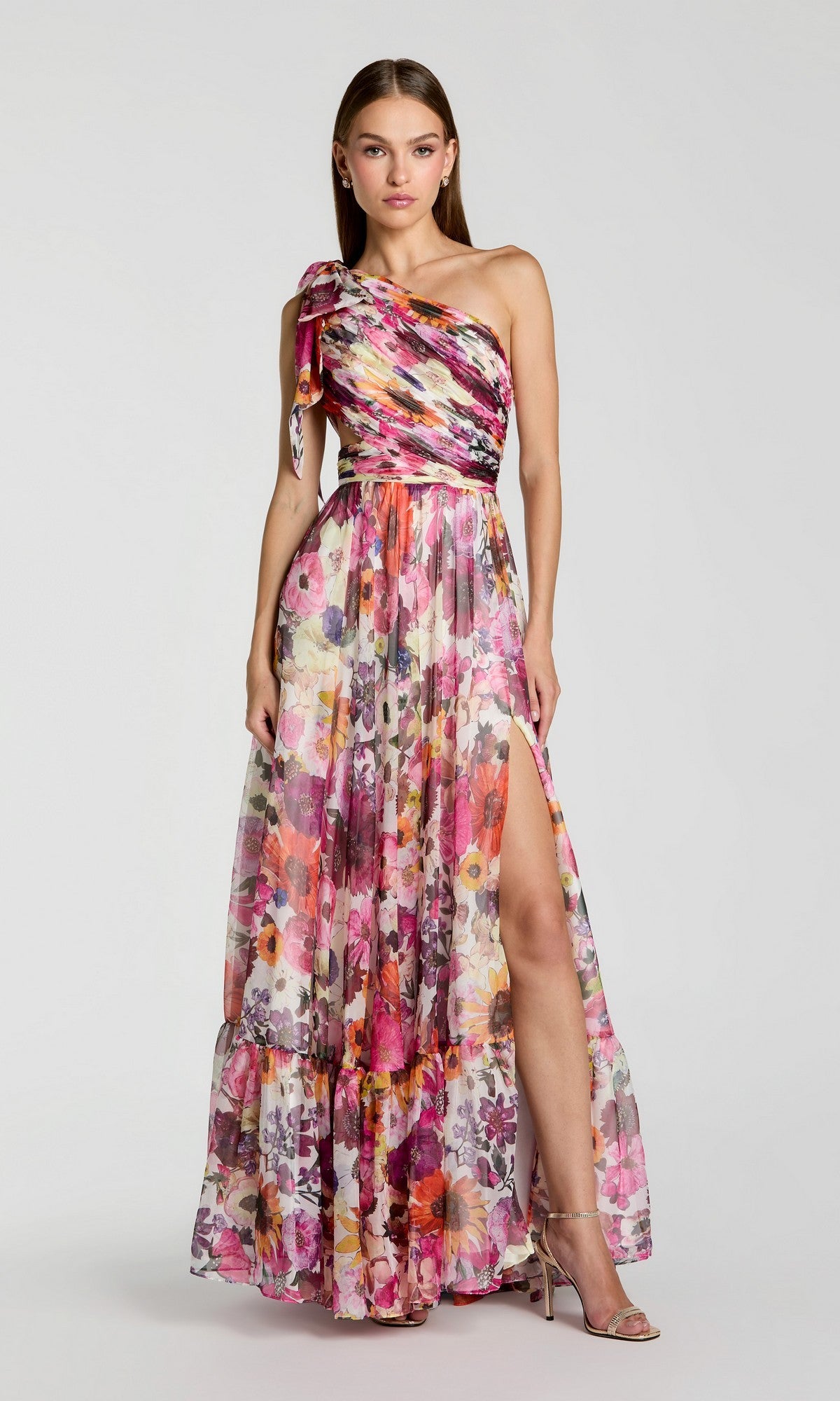 A woman stands in heels wearing the Mac Duggal 68553 long formal dress in White Multi—a chic, one-shoulder A-line gown with floral print, soft chiffon fabric, floor length, a side slit, and elegant sophistication against a plain background.